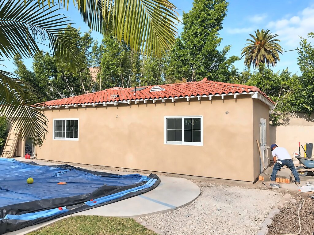 Detached garage in Los Angeles converted to an ADU, with new stucco, a red tile roof and white windows, beside a covered pool