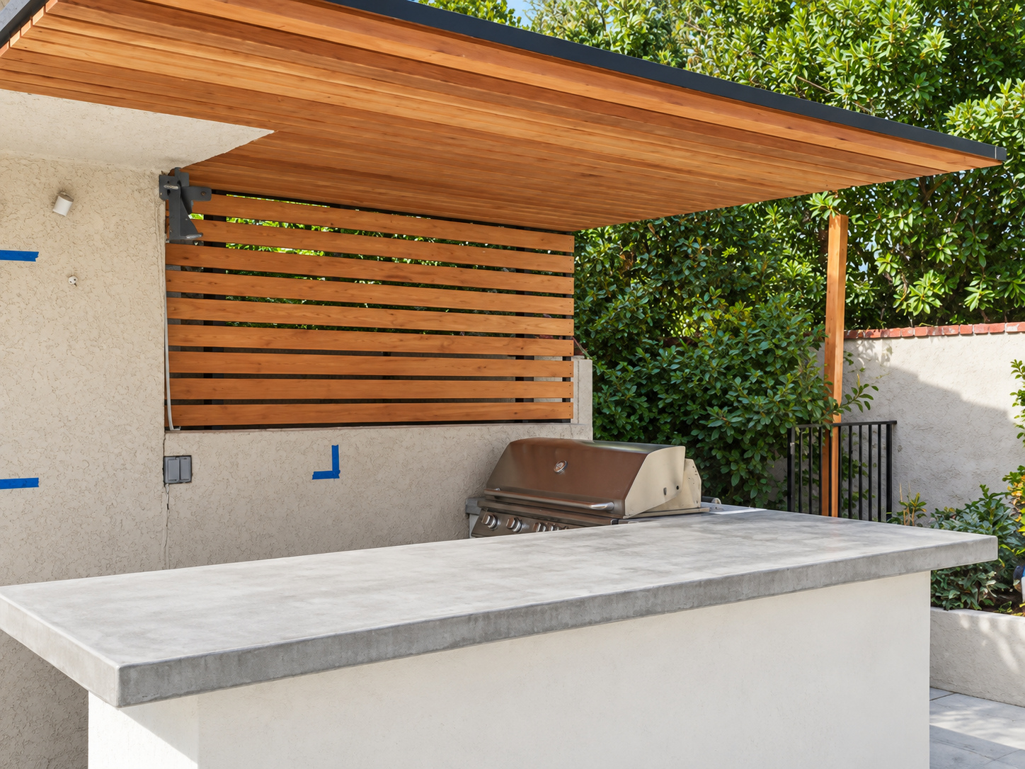 Outdoor kitchen with a poured concrete island, built-in grill and cantilevered cedar slat cover over travertine paving, built by Green Design and Build