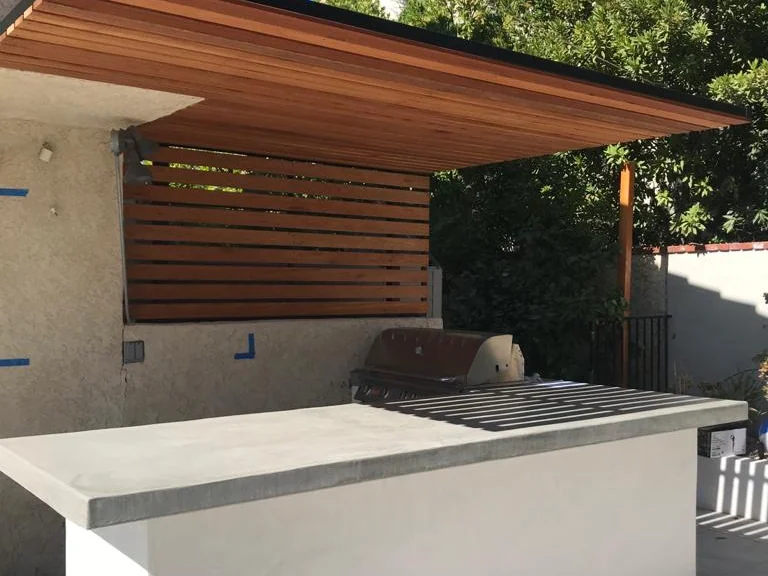 Outdoor kitchen with a poured concrete island, built-in grill and cantilevered cedar slat cover over travertine paving, built by Green Design and Build